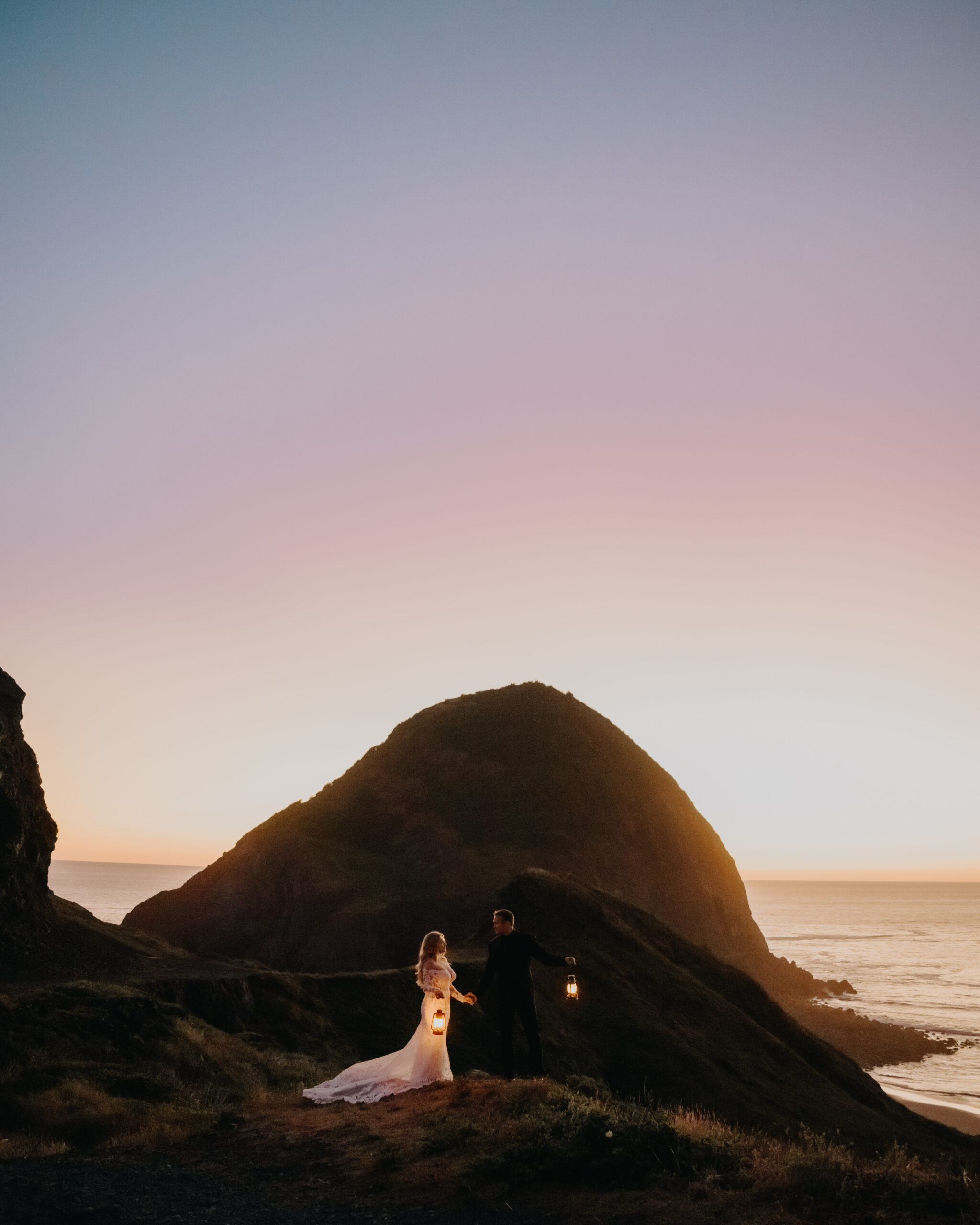 Bride and groom walk along Oregon coast with lanterns in hand at dusk