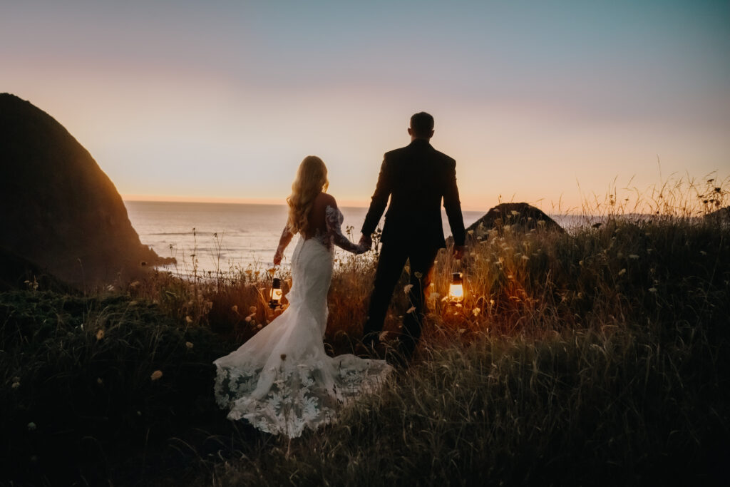 A bride and groom walk towards the ocean on the coast of Oregon during their adventurous destination elopement, holding lanterns to light their path at dusk, with little light remaining from the sun setting minutes before.