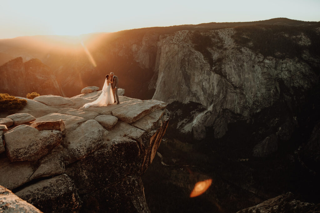 Bride and groom share an intimate moment at Taft Point in Yosemite National Park during their destination elopement, standing on the edge of a dramatic cliff with sweeping views of Yosemite Valley and El Capitan in the distance. The bride's dress flows in the mountain breeze as golden hour sunlight covers the cliffs, captured by The Pinckards, expert Yosemite elopement photographers and videographers specializing in adventurous, scenic weddings.