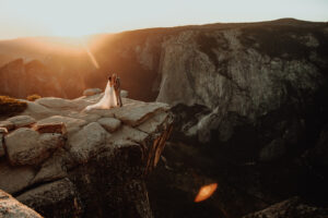 Bride and groom share an intimate moment at Taft Point in Yosemite National Park during their destination elopement, standing on the edge of a dramatic cliff with sweeping views of Yosemite Valley and El Capitan in the distance. The bride's dress flows in the mountain breeze as golden hour sunlight covers the cliffs, captured by The Pinckards, expert Yosemite elopement photographers and videographers specializing in adventurous, scenic weddings.