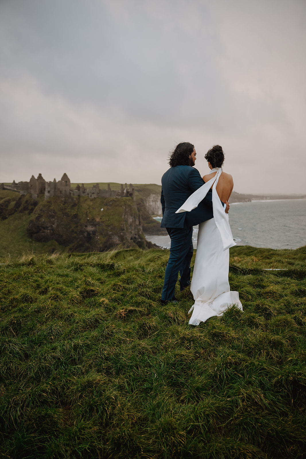 Bride and groom eloping on the cliffs of Northern Ireland with Dunluce Castle ruins in the background, captured during a windswept coastal destination elopement overlooking the Atlantic Ocean. Perfect for couples planning an adventurous and intimate Ireland elopement experience with cinematic photography and videography.