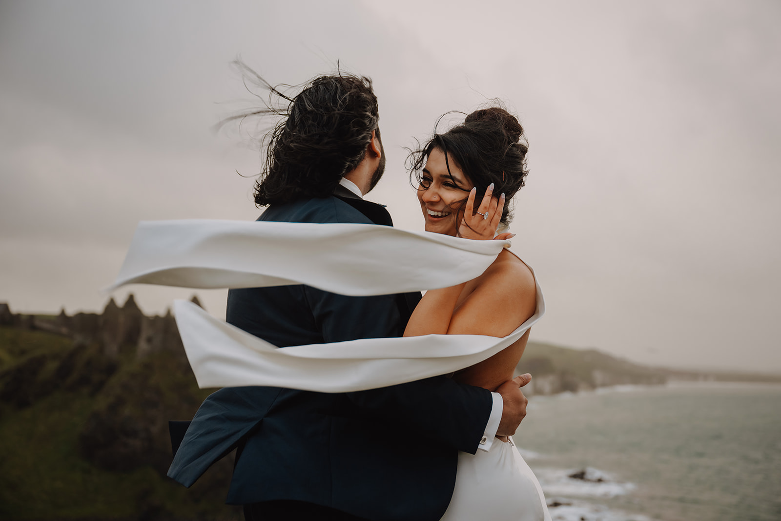 Bride and groom embrace as the wind blows their hair and her dress standing over the coast of Northern Ireland with Dunluce Castle in the background