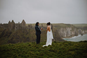 Bride and groom reading vows to each other at the edge of coastal cliffs in northern Ireland with the Dunluce Castle in the background