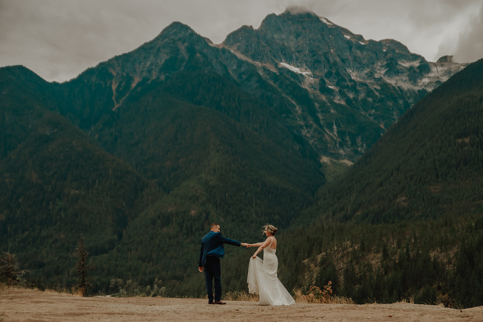 Bride and groom stand on a rugged cliffside at sunset during their adventurous destination elopement in the North Cascades National Park, with the golden light from sunset casting a romantic glow over the dramatic landscape. The bride wears a flowing lace gown, and the groom embraces her from behind as they overlook a sweeping valley view, captured by The Pinckards, destination elopement photographers and videographers specializing in intimate, nature-filled weddings.