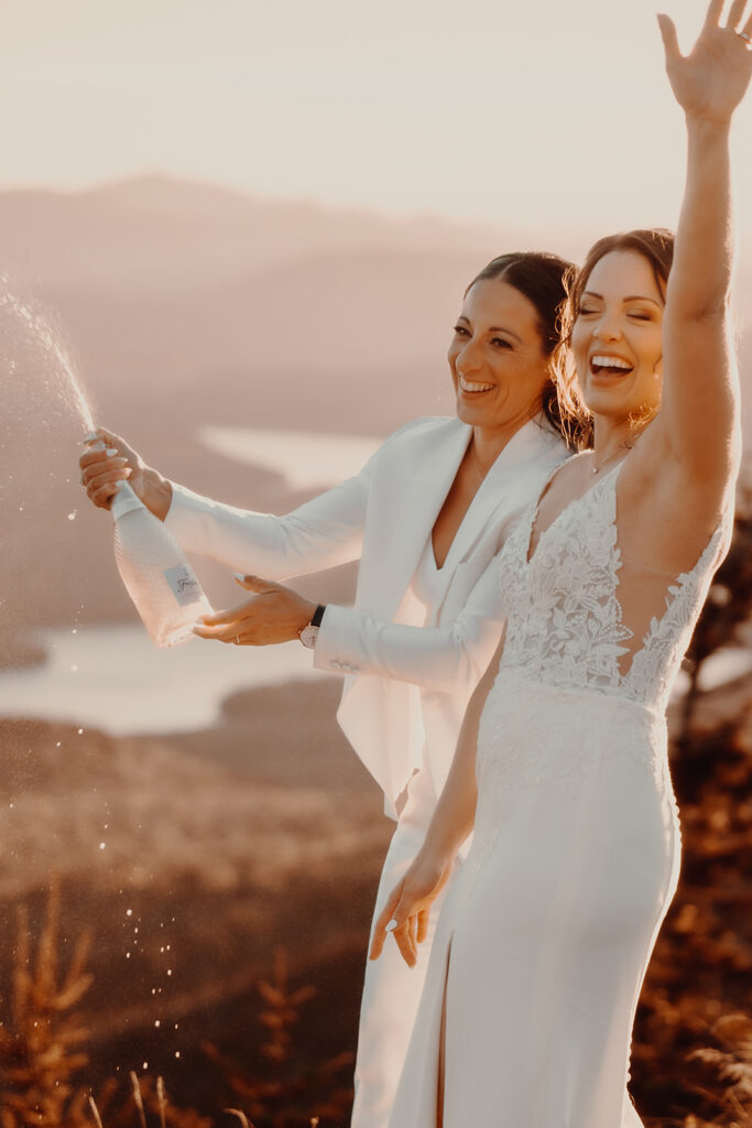 Two brides popping champagne on top of Whiteface Mountain in Lake Placid, NY during their sunset elopement ceremony